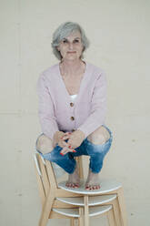 Lächelnde ältere Frau mit grauen Haaren, die auf Stühlen vor einer Wand hockt - JOSEF09384