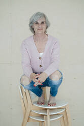 Smiling senior woman with gray hair crouching on chairs in front of wall - JOSEF09384