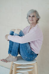 Smiling senior woman with gray hair sitting on chairs in front of wall - JOSEF09385