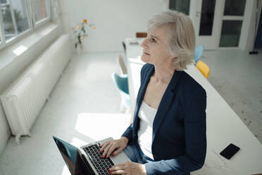 Senior businesswoman with laptop sitting on desk in office - JOSEF09390