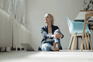 Senior businesswoman with laptop sitting on ground in office - JOSEF09392