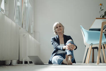 Lächelnde ältere Geschäftsfrau mit Laptop auf dem Boden sitzend im Büro - JOSEF09393