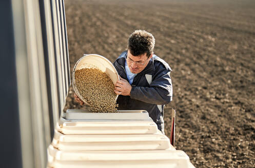 Männlicher Landwirt gießt Sojabohnensamen in eine Maschine auf einem Feld - NOF00500