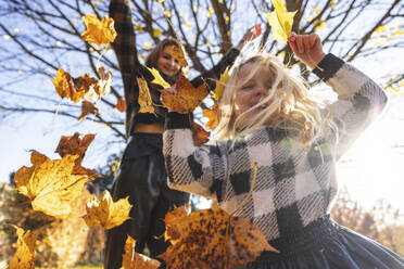 Mother throwing autumn leaves on daughter playing at park - WPEF05959