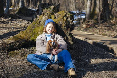 Smiling young woman with pet dog sitting in forest on sunny day - OMIF00781