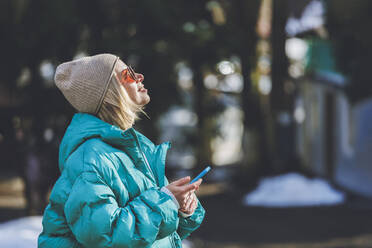 Smiling woman wearing sunglasses holding smart phone on sunny day - OMIF00789