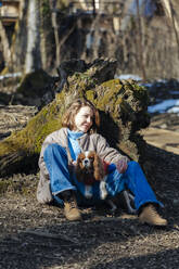 Smiling young woman with dog sitting in forest on sunny day - OMIF00792