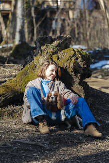 Lächelnde junge Frau mit Hund sitzt im Wald an einem sonnigen Tag - OMIF00792