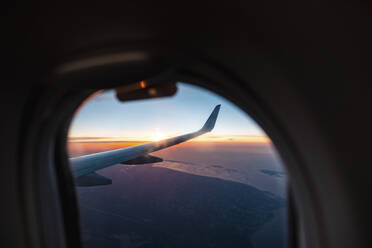Wing of airplane flying against setting sun seen through porthole - WPEF05965