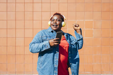 Woman holding smart phone listening music through wireless headphones in front of wall - MEUF05479