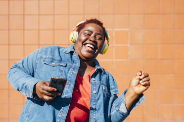 Fröhliche Frau mit Smartphone, die über drahtlose Kopfhörer Musik hört und sich vor einer Wand amüsiert - MEUF05480