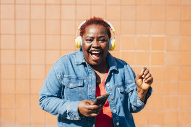 Fröhliche Frau mit Smartphone vor einer Wand, die Musik genießt - MEUF05481