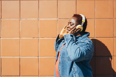 Woman enjoying music through wireless headphones in front of wall - MEUF05482