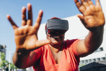 Glückliche Frau in einem Virtual-Reality-Simulator gestikuliert an einem sonnigen Tag - MEUF05492