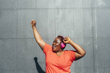 Happy woman enjoying music dancing with hand raised in front of wall - MEUF05496