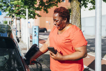 Glückliche Frau mit Handy in der Hand, die an einem sonnigen Tag ein Elektroauto auflädt - MEUF05508