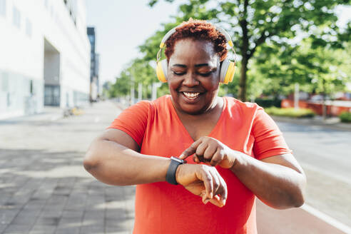Glückliche Frau, die Musik über drahtlose Kopfhörer hört und ihre Smartwatch kontrolliert - MEUF05518