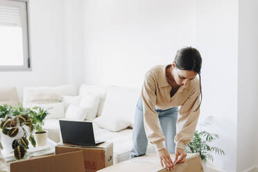 Young woman unpacking cardboard box in living room at home - MRRF02078
