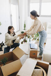 Woman giving potted plant to girlfriend unpacking in living room at home - MRRF02081