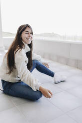 Glückliche junge Frau mit langen Haaren sitzt auf der Terrasse - MRRF02086