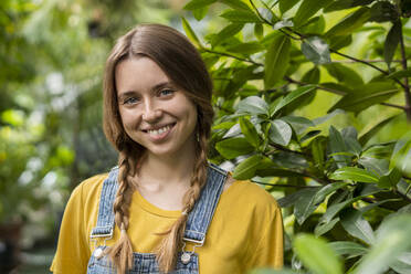 Lächelnde junge Frau steht bei einer Pflanze im Garten - SSGF00856