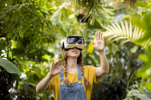 Young woman wearing virtual reality simulator gesturing in garden - SSGF00869