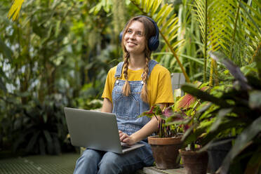 Glückliche Frau mit Laptop hört Musik über drahtlose Kopfhörer im Garten - SSGF00872