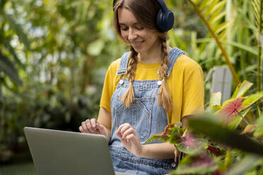 Glückliche junge Frau mit drahtlosen Kopfhörern, die mit einem Laptop im Garten sitzt - SSGF00874