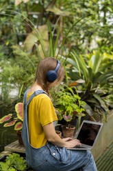 Junge Frau mit Laptop, die im Garten sitzend über drahtlose Kopfhörer Musik hört - SSGF00876
