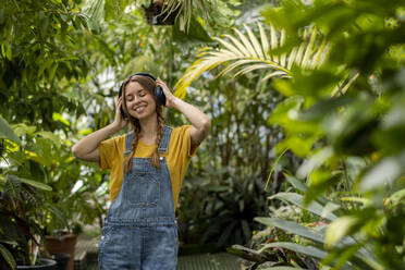 Glückliche Frau mit geschlossenen Augen, die im Garten Musik über drahtlose Kopfhörer hört - SSGF00879
