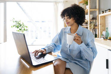 Smiling woman holding coffee cup using laptop sitting at kitchen island - OIPF01688