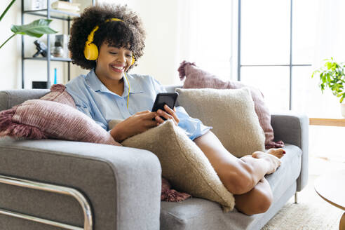Glückliche junge Frau mit Kopfhörern und Smartphone auf dem Sofa zu Hause - OIPF01699