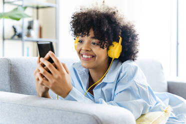 Happy woman with headphones using smart phone lying on sofa at home - OIPF01701