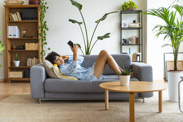 Smiling woman taking selfie through smart phone lying on sofa at home - OIPF01704