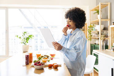 Junge Frau benutzt einen Tablet-PC und isst eine Erdbeere in der Küche zu Hause - OIPF01712