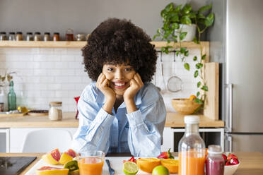 Lächelnde schöne Frau mit Afro-Frisur lehnt sich durch Früchte auf Kücheninsel zu Hause - OIPF01724