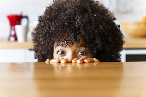 Woman with Afro hairstyle hiding behind kitchen island at home - OIPF01727