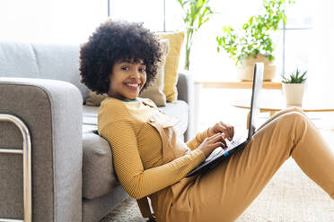Smiling woman with laptop leaning on sofa at home - OIPF01738