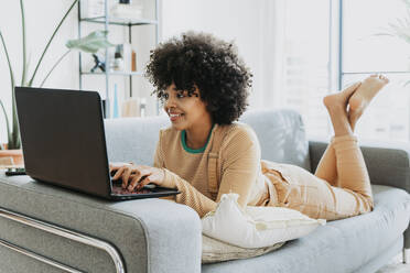 Smiling woman using laptop lying on sofa at home - OIPF01740