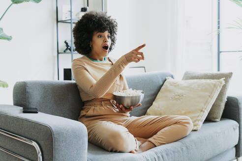 Shocked Afro woman watching TV sitting on sofa with bowl of popcorn pointing at home - OIPF01745