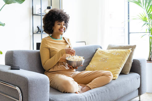Happy woman holding popcorn bowl sitting on sofa - OIPF01746
