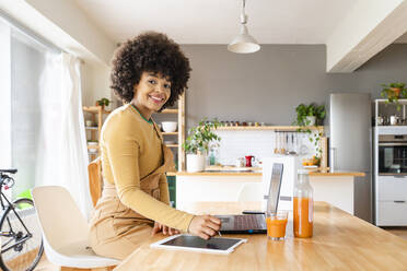 Glückliche junge Frau mit Tablet-PC und Laptop am Tisch im Wohnzimmer sitzend - OIPF01757