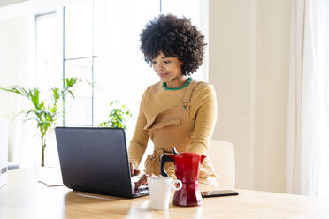 Young woman using laptop by coffee pot and cup on table at home - OIPF01758