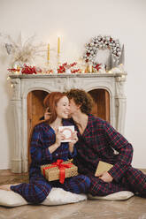 Man kissing girlfriend holding greeting card sitting with gift in front of fireplace at home - EIF04093