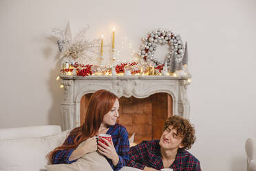 Smiling couple holding cup sitting in front of fireplace at home - EIF04096