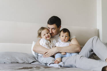 Smiling man embracing son and daughter sitting on bed at home - SEAF00864