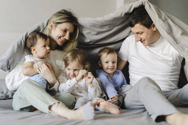 Happy parents with children sitting under blanket in bedroom - SEAF00866