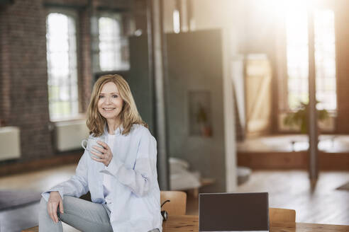 Lächelnde Frau mit Kaffeetasse, die zu Hause auf dem Tisch sitzt - FMKF07562