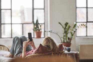 Frau mit Smartphone auf dem Sofa liegend zu Hause - FMKF07567