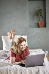 Smiling woman using laptop lying on bed at home - FMKF07574